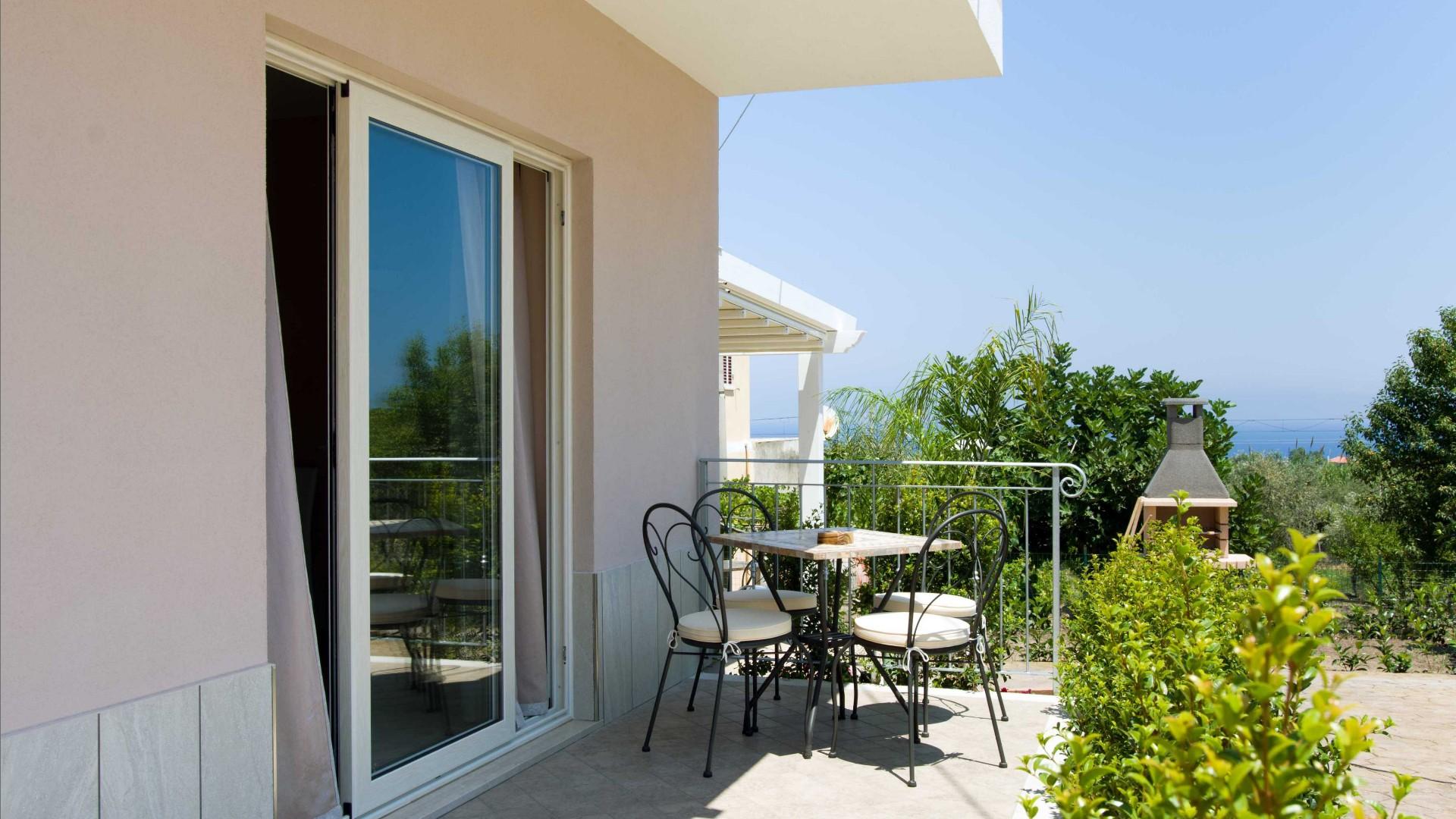 Terrazza con tavolo, sedie e barbecue, circondata da vegetazione e vista sul mare.