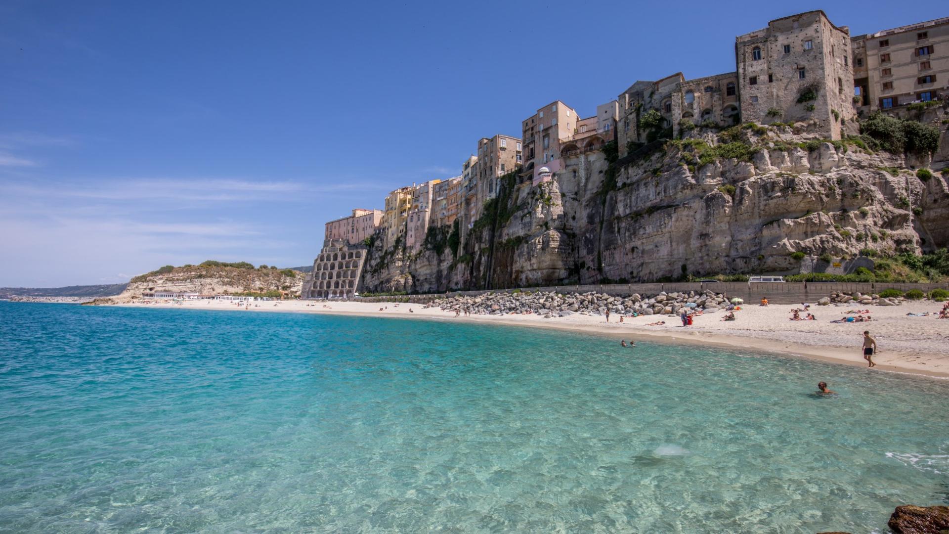 Spiaggia con acqua cristallina e case su scogliera, sotto un cielo sereno.