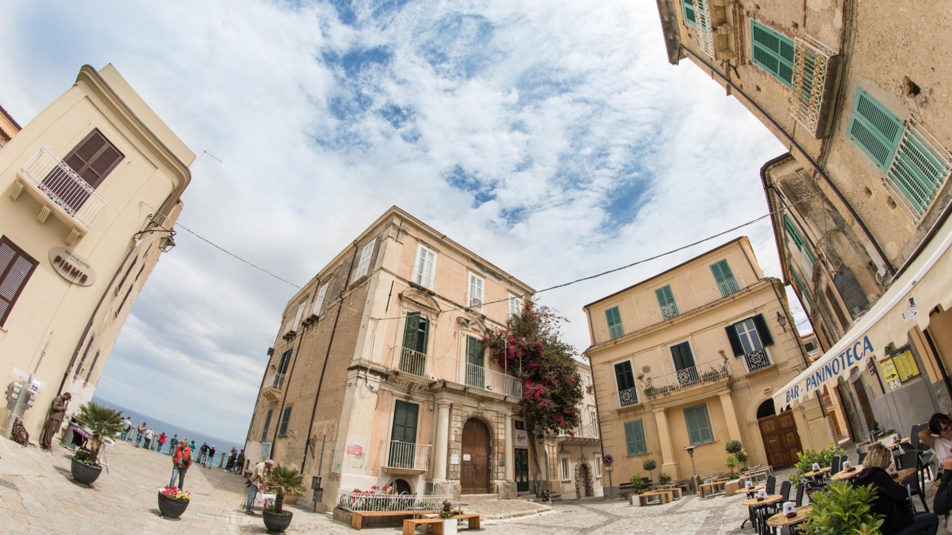 Piazza pittoresca con edifici storici, caffè all'aperto e vista sul mare.