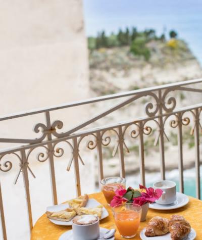 Colazione con vista mare: cappuccino, succo, croissant e fiori su un tavolo giallo.