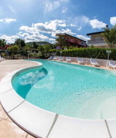 Piscina all'aperto con sdraio, cielo sereno e case sullo sfondo.
