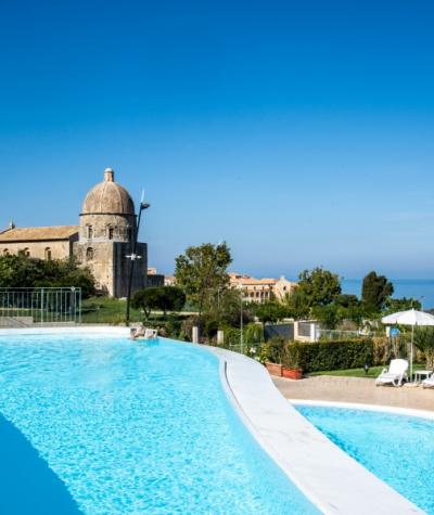 Piscina con vista mare e antica chiesa, cielo limpido e sedie a sdraio.